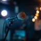 A close-up of a microphone on a stand with a blurred background of stage lights.